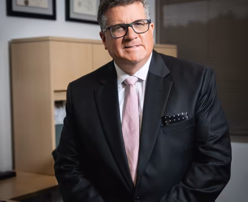 Image of Christopher Finney sitting on his desk, black suit, pink tie and polka dot pocket square