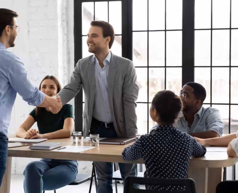 Five young professionals at a maple table with two men standing and shaking hands